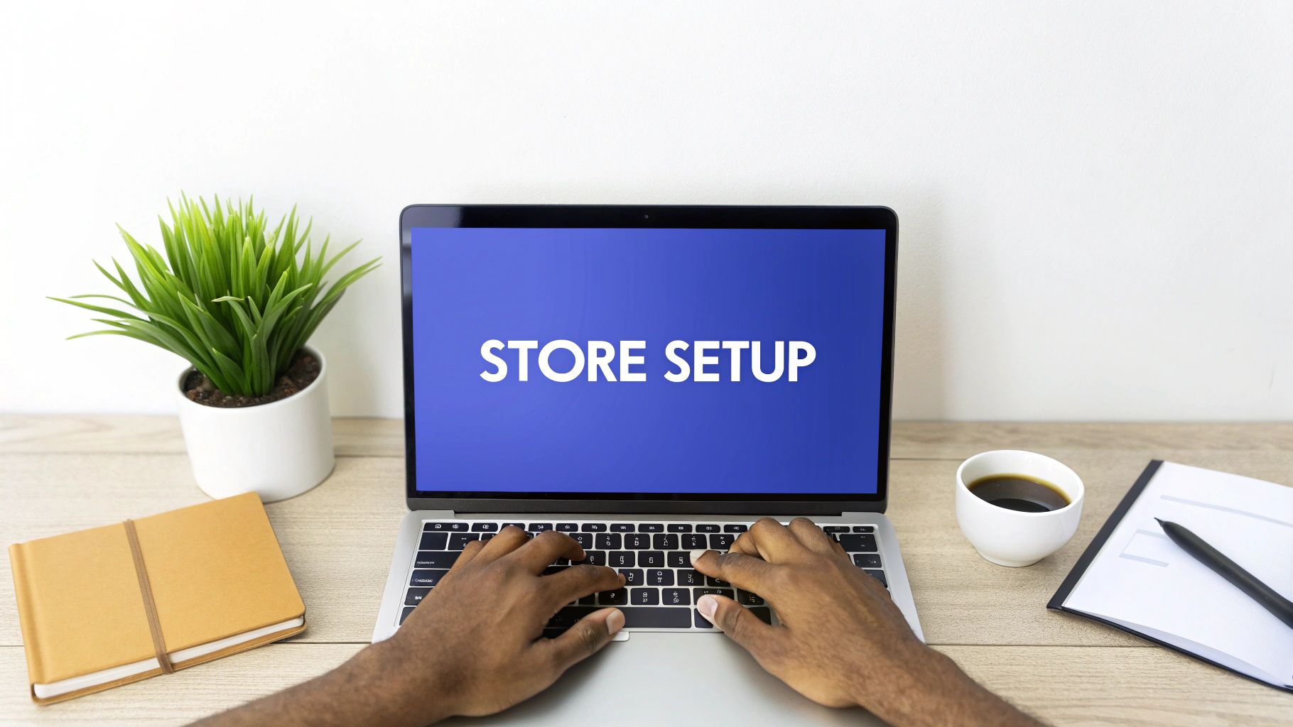 A person types on a laptop displaying ‘STORE SETUP’, surrounded by a plant, coffee, and notebook.